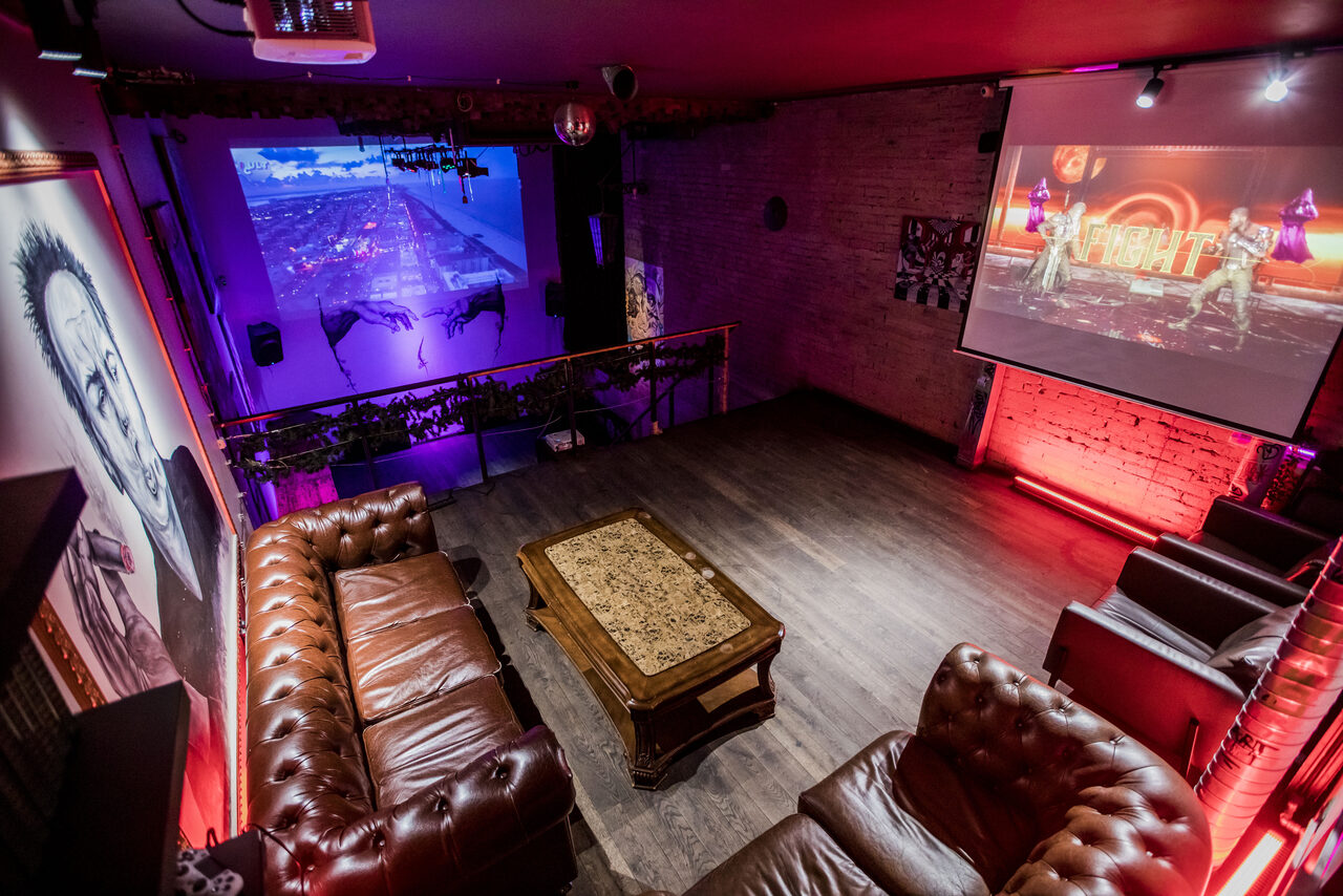 Арендовать Street Art Loft (Стрит Арт Лофт). Арт-пространство с диванами и журнальным столиком, проектор и экран с изображением, второй экран на стене. Кирпичные стены, деревянный пол, мягкая мебель из коричневой кожи. Красная неоновая подсветка, подвесные светильники, настенные картины.