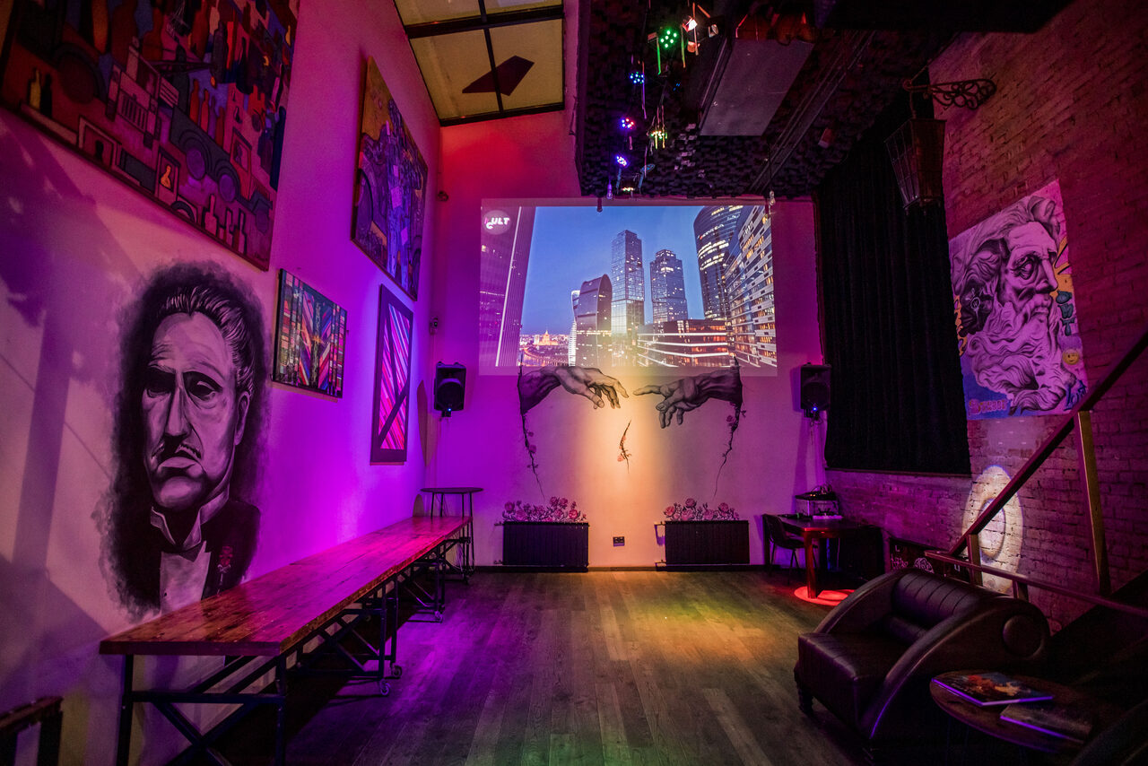 Арендовать Street Art Loft (Стрит Арт Лофт). Арт-пространство с кирпичными стенами, украшенное картинами и граффити. Длинная деревянная скамья, кожаный диван, проектор с изображением города. Фиолетовая и розовая подсветка, темный деревянный пол.