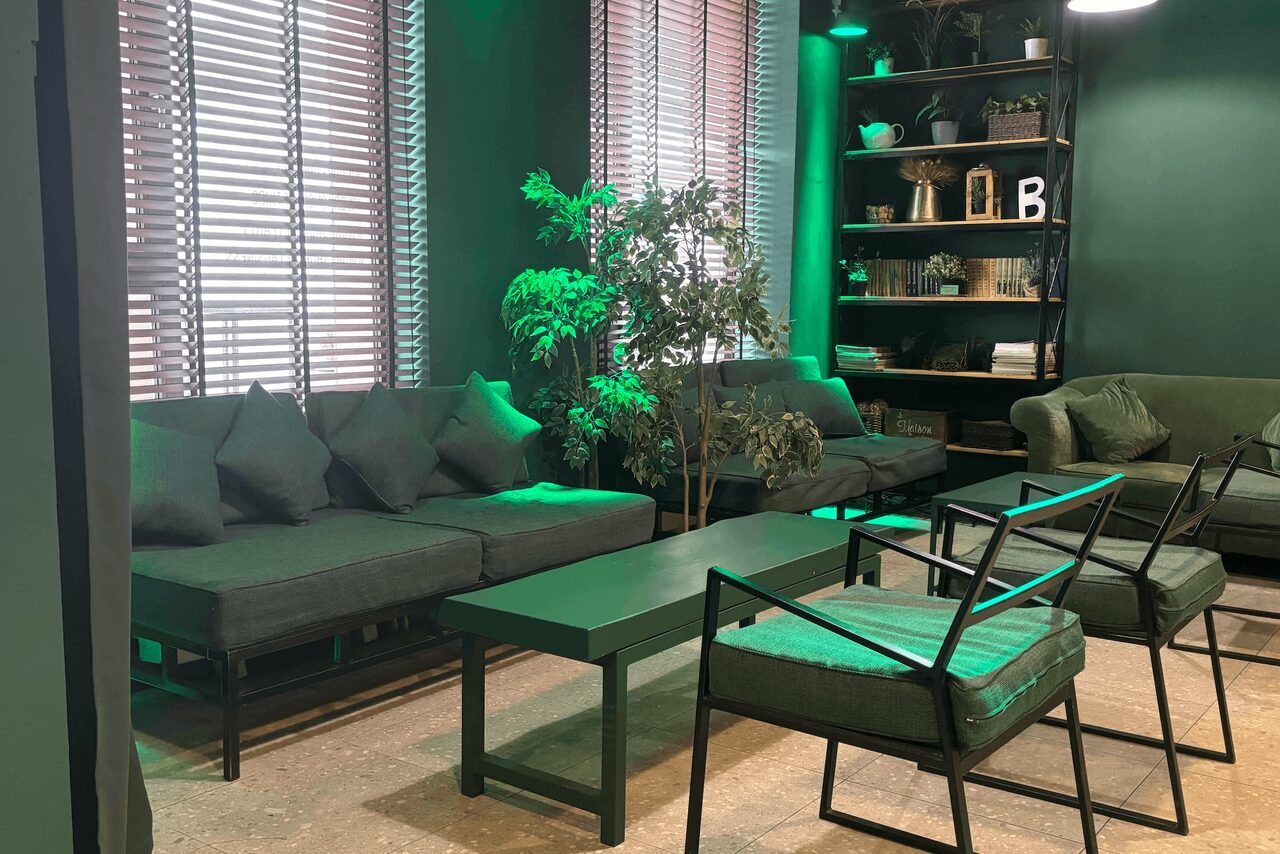 Арендовать Work Shop Loft(Воркшоп Лофт). Зал 11 . Современная гостиная в зелёных тонах с диванами и креслами, кофейным столиком, стеллажом с декором. На полу светлая поверхность, стены темно-зелёные, окна с жалюзи, комнатные растения добавляют уюта.