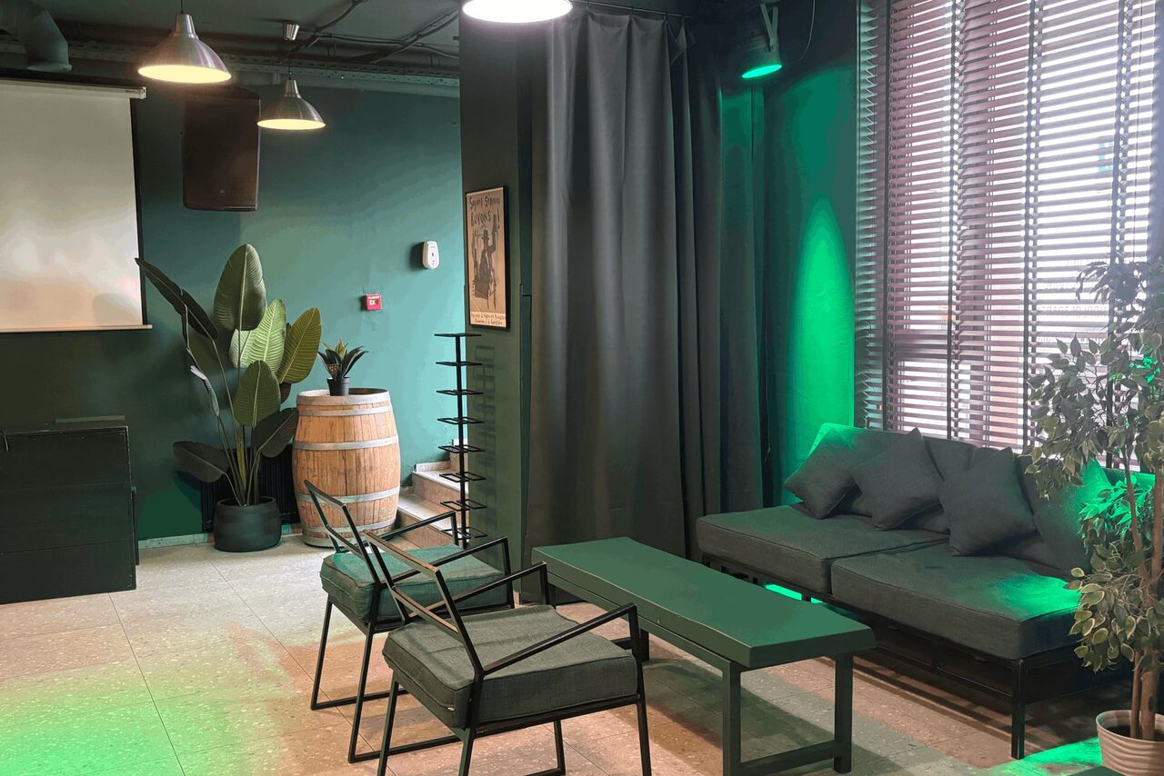 Арендовать Work Shop Loft(Воркшоп Лофт). Зал 11 . Пространство с диваном и креслами у окна с жалюзи, металлический стол, зеленая подсветка, черные шторы, бочонок с растением, полки, проектор, подвесные светильники, зеленые стены, декоративное растение.