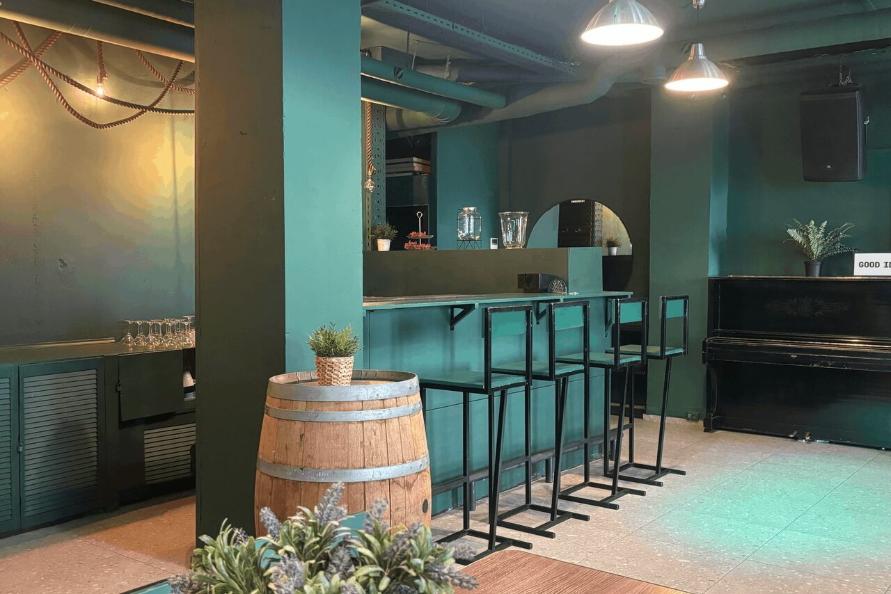 Арендовать Work Shop Loft(Воркшоп Лофт). Зал 11 . Барное пространство с высокими табуретами, стойкой и деревянной бочкой. Стены и мебель зелёного цвета. Справа чёрное пианино. Подвесные светильники. Растения в горшках. Плиточный пол светлого оттенка. Слева столы у стены.