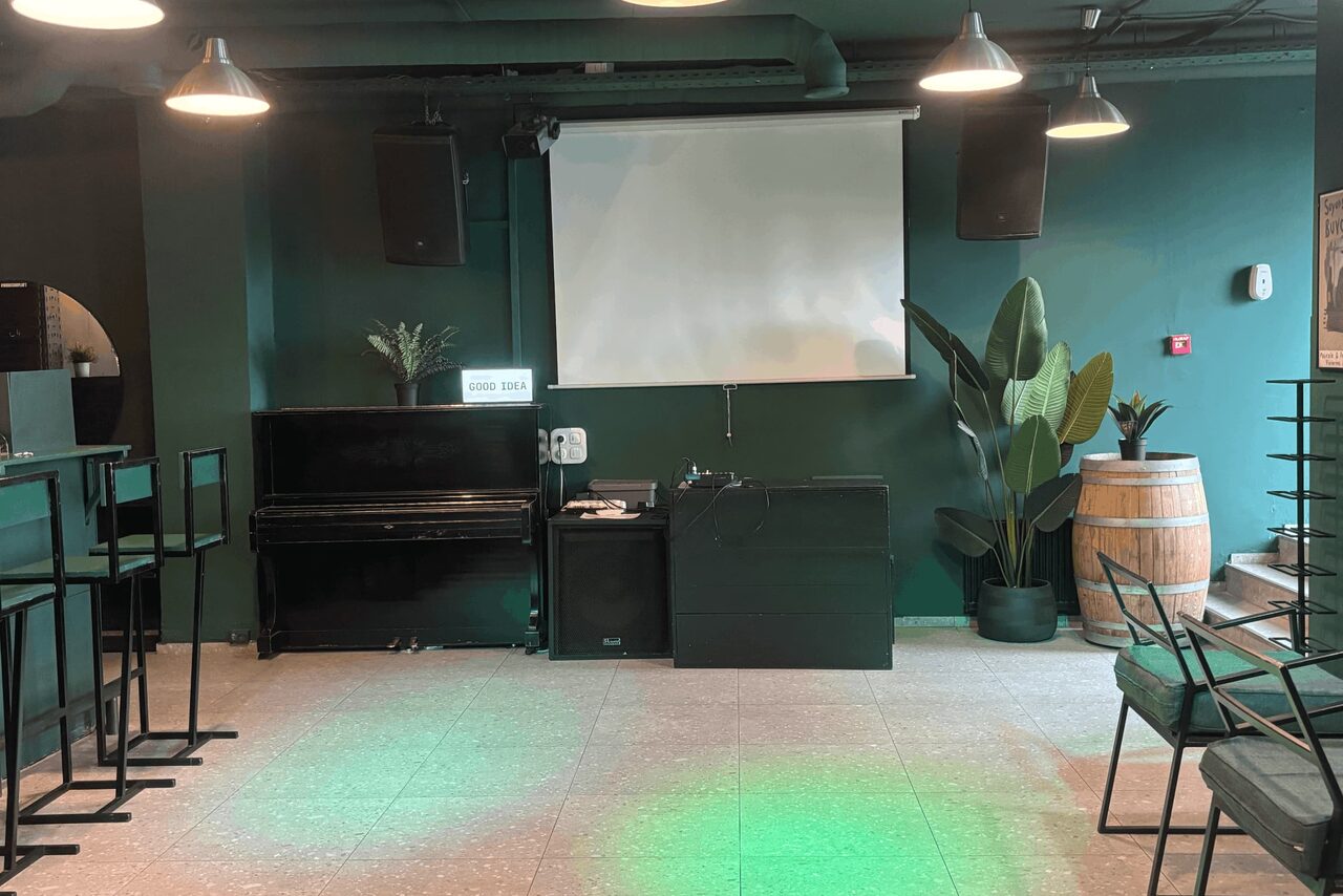 Арендовать Work Shop Loft(Воркшоп Лофт). Зал 11 . Бар с темно-зелеными стенами, столами и стульями в индустриальном стиле. На стене развешаны два громкоговорителя и большой экран. Справа - деревянная бочка и растения. Потолочные светильники создают мягкое освещение. На полу плитка.