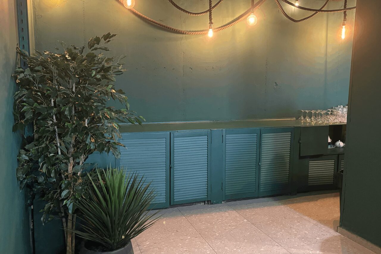 Арендовать Work Shop Loft(Воркшоп Лофт). Зал 11 . Небольшое помещение с зелеными стенами и плиточным полом. Вдоль стены расположен стол с зелеными дверцами. На столе стоит ряд бокалов. Над головой висят лампы в виде оголенных лампочек на шнурах. В углу расположены искусственные растения.