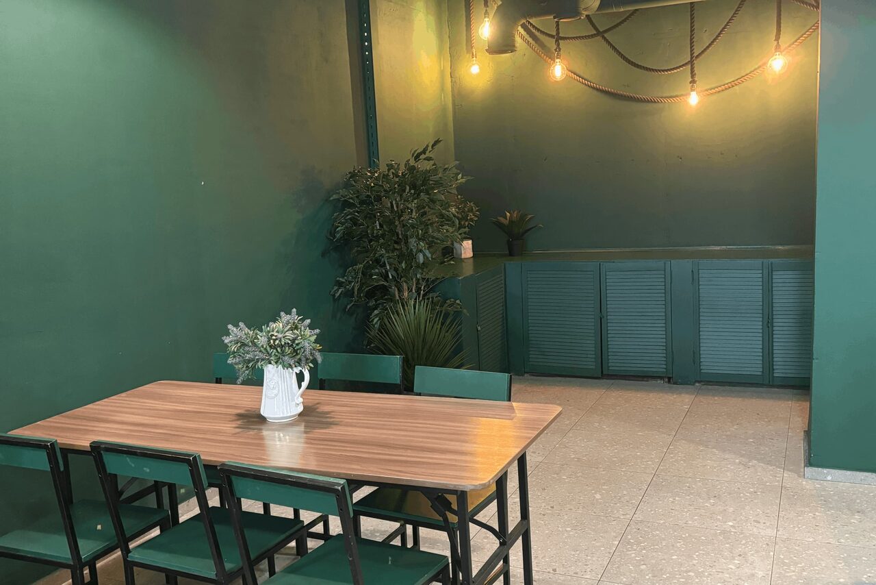 Арендовать Work Shop Loft(Воркшоп Лофт). Зал 11 . Пространство с зелеными стенами и потолком, деревянный стол с шестью стульями, декор из растений, подвесные лампы на веревках, закрытые шкафы, светло-серый плиточный пол, простое расположение.