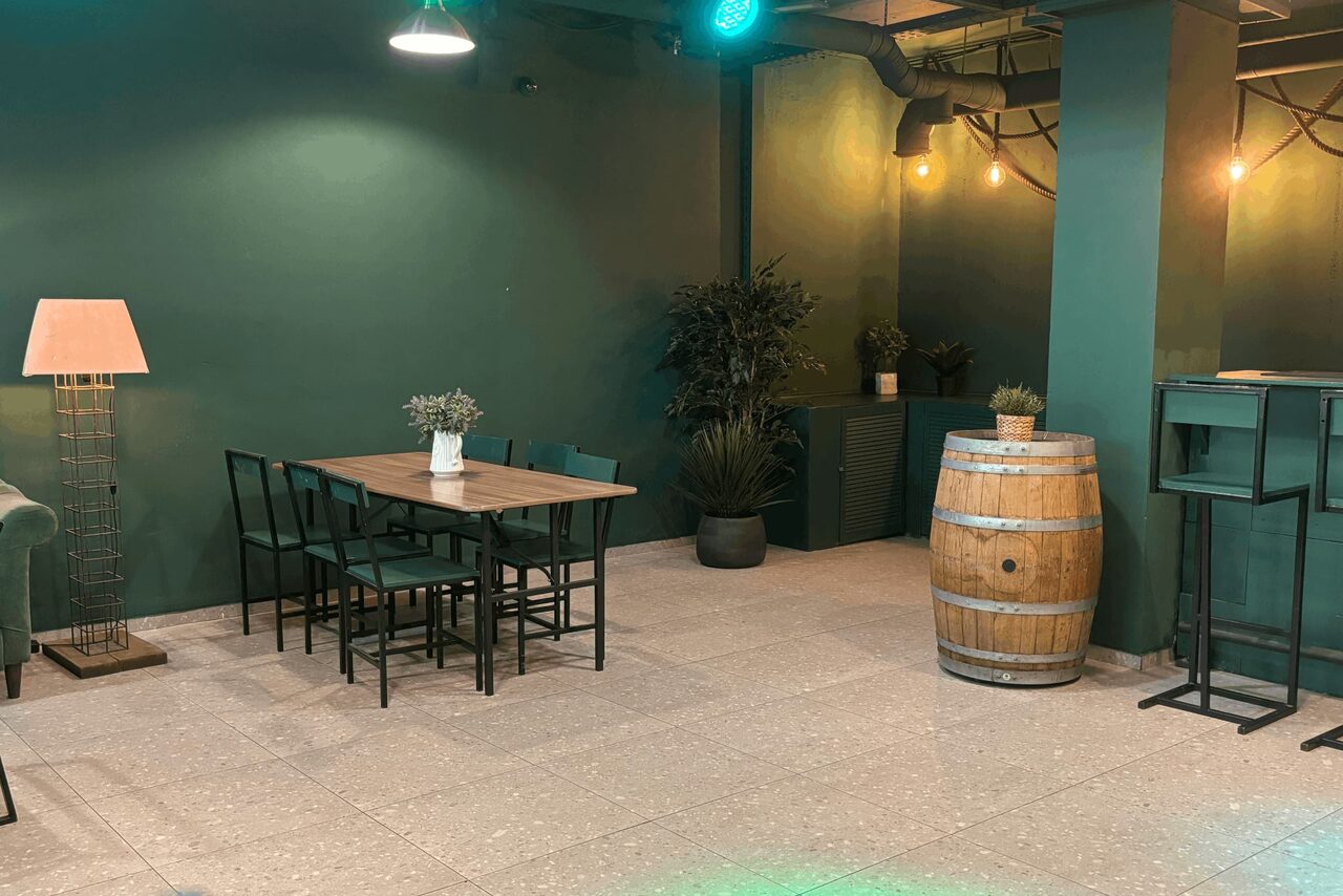 Арендовать Work Shop Loft(Воркшоп Лофт). Зал 11 . Пространство с зелеными стенами и терракотовым полом. Деревянный стол с восемью стульями, рядом бочка и высокая лампа. В углу растения и металлический шкаф. Потолочные светильники и лампочки на стенах.