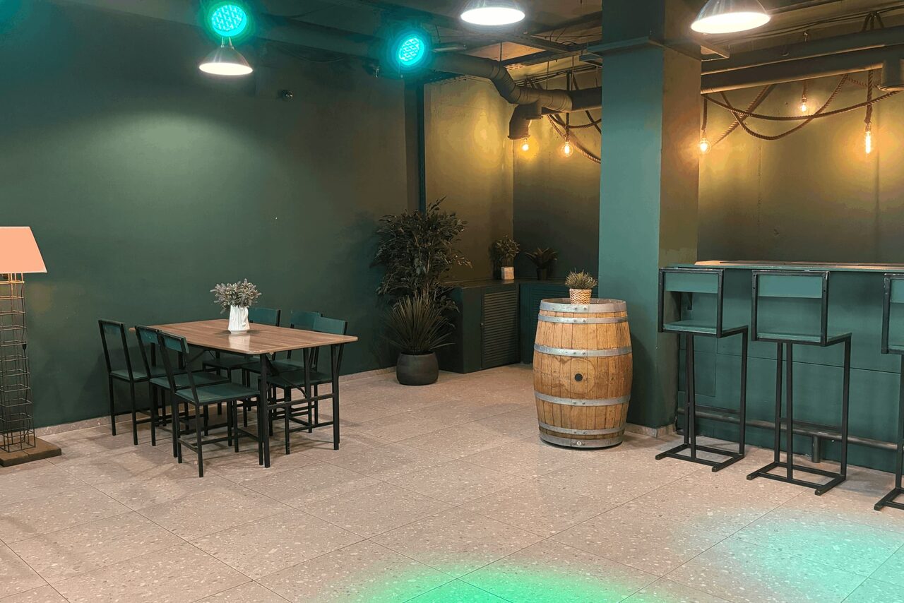 Арендовать Work Shop Loft(Воркшоп Лофт). Зал 11 . Мультфункциональное пространство с зелеными стенами, столом и стульями слева, барной стойкой и стульями справа. Напольное покрытие светлое плиточное. На стенах декоративные светильники, вокруг зеленые растения в горшках.