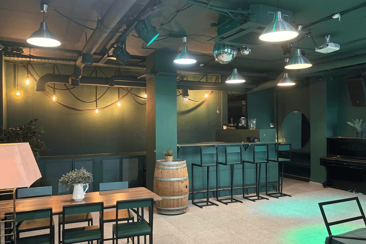 Арендовать Work Shop Loft(Воркшоп Лофт). Зал 11 . Многофункциональное пространство с барной стойкой, высокими стульями и деревянными столами. На стене винтажные лампы, подвесные светильники на потолке, пианино в углу. Потолочные балки, зелёные стены, бочка-стол, панорамные окна.
