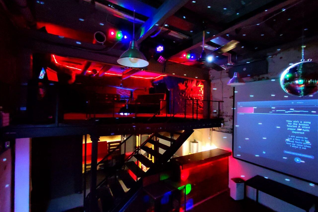 Арендовать Soul Loft . Затемнённая комната лофта, много людей в кадре проводят фуршетное мероприятие, беря еду с объединённых столов. Несколько людей на кадре танцуют