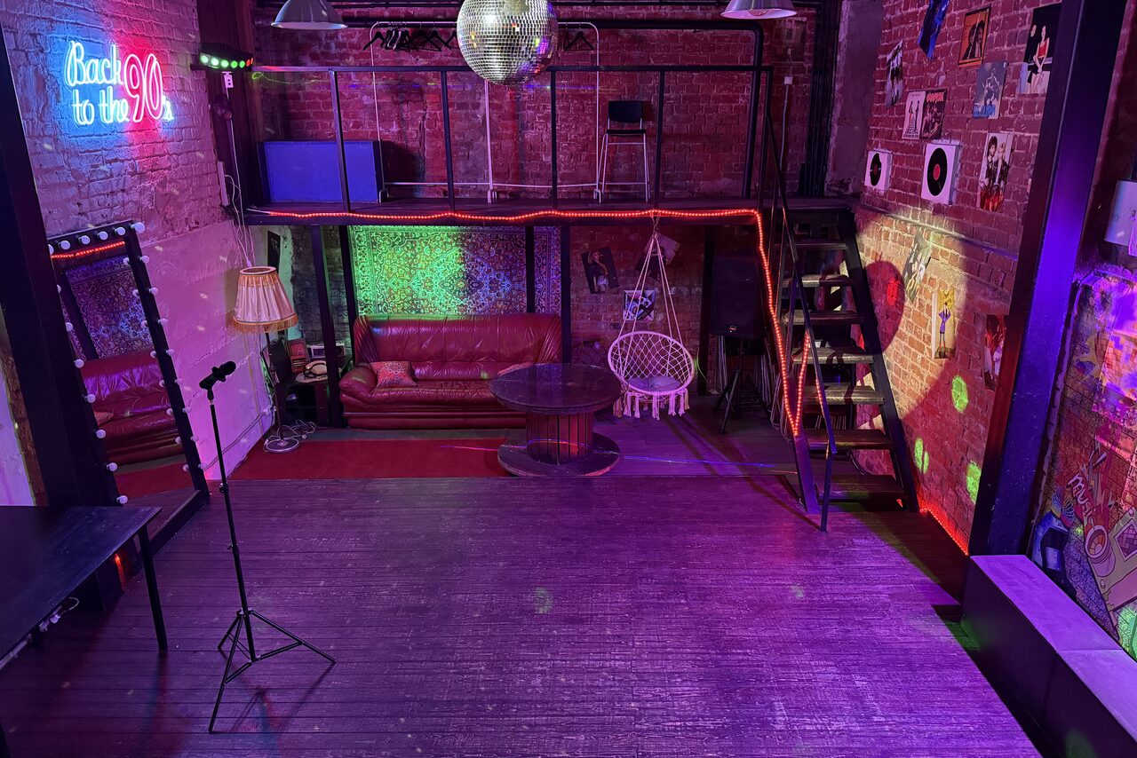 Арендовать Лофт ИЗНАНКА. Зал ЛИХИЕ 90-е. Затемнённая комната лофта, много людей в кадре проводят фуршетное мероприятие, беря еду с объединённых столов. Несколько людей на кадре танцуют