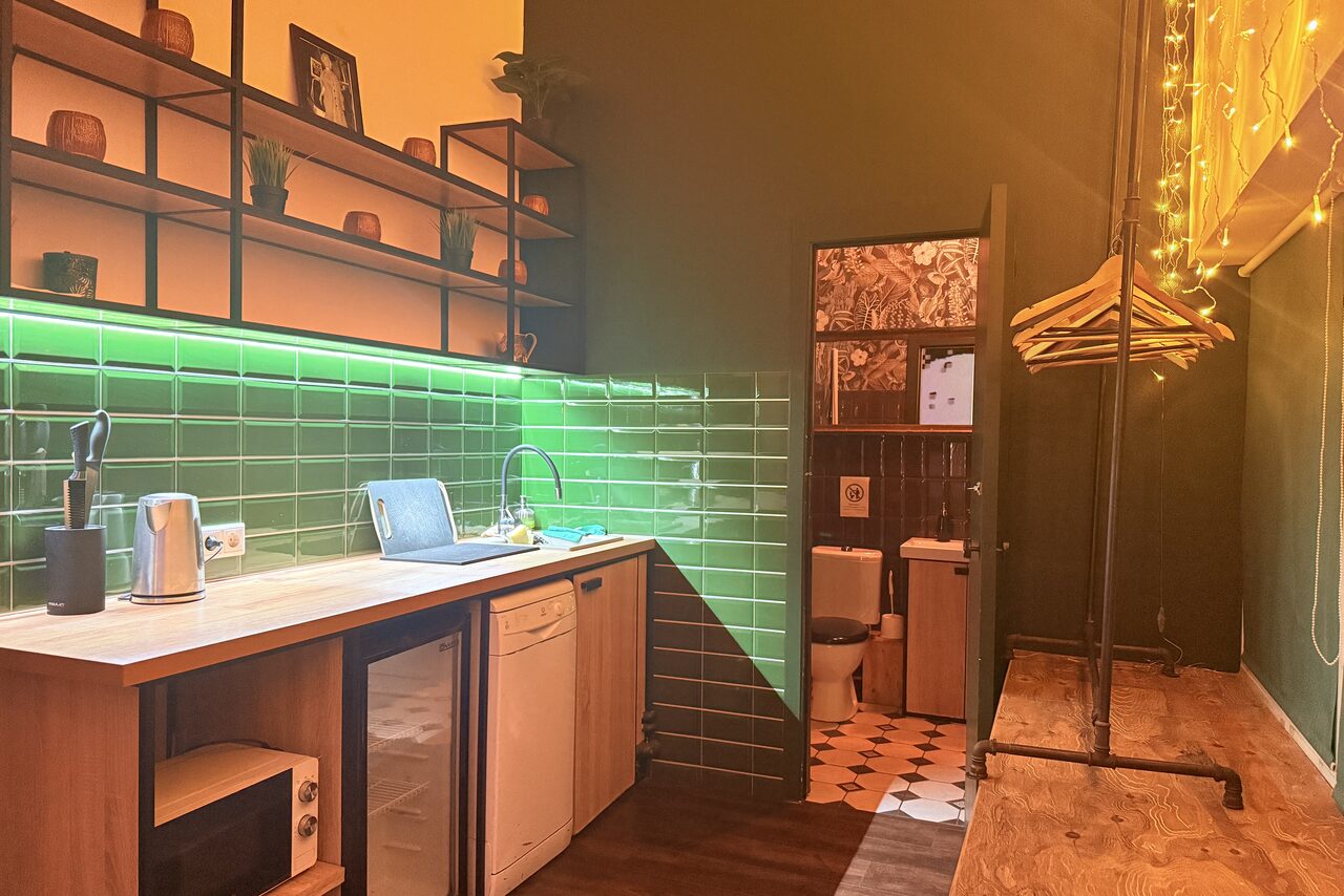 Арендовать Bar on Presnya for private events. Кухонное пространство с деревянной столешницей, мойкой и посудомоечной машиной. Зелёная плитка на стене с подсветкой. Полка с декором. Открытая вешалка с вешалками. Видна часть туалета с плиточным полом. Тёплое освещение.