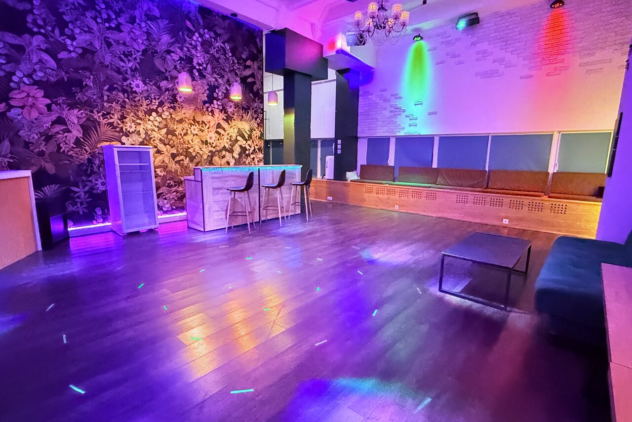 Арендовать Bar on Presnya for private events. Просторное помещение с деревянным полом, барной стойкой и высокими стульями, диванами вдоль стены. Освещение разноцветное, с акцентом на стенах. Темные обои с растительным узором. Потолок украшен люстрой.