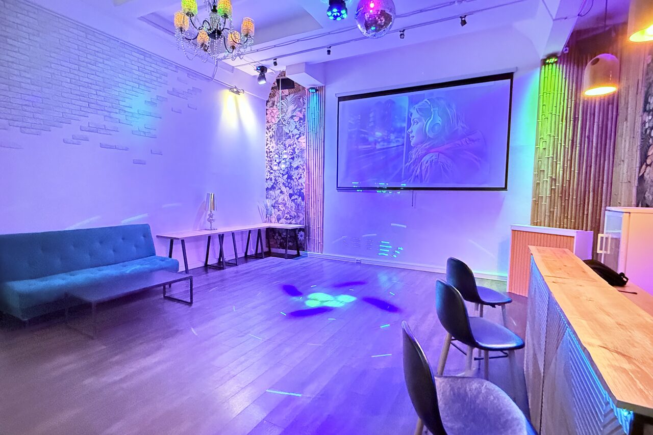 Арендовать Bar on Presnya for private events. Современное просторное помещение с белыми кирпичными стенами и деревянным полом. Мягкий синий диван и несколько стульев вокруг деревянного стола. Настенные декорации, проектор с экраном, разноцветные светильники и барная стойка.