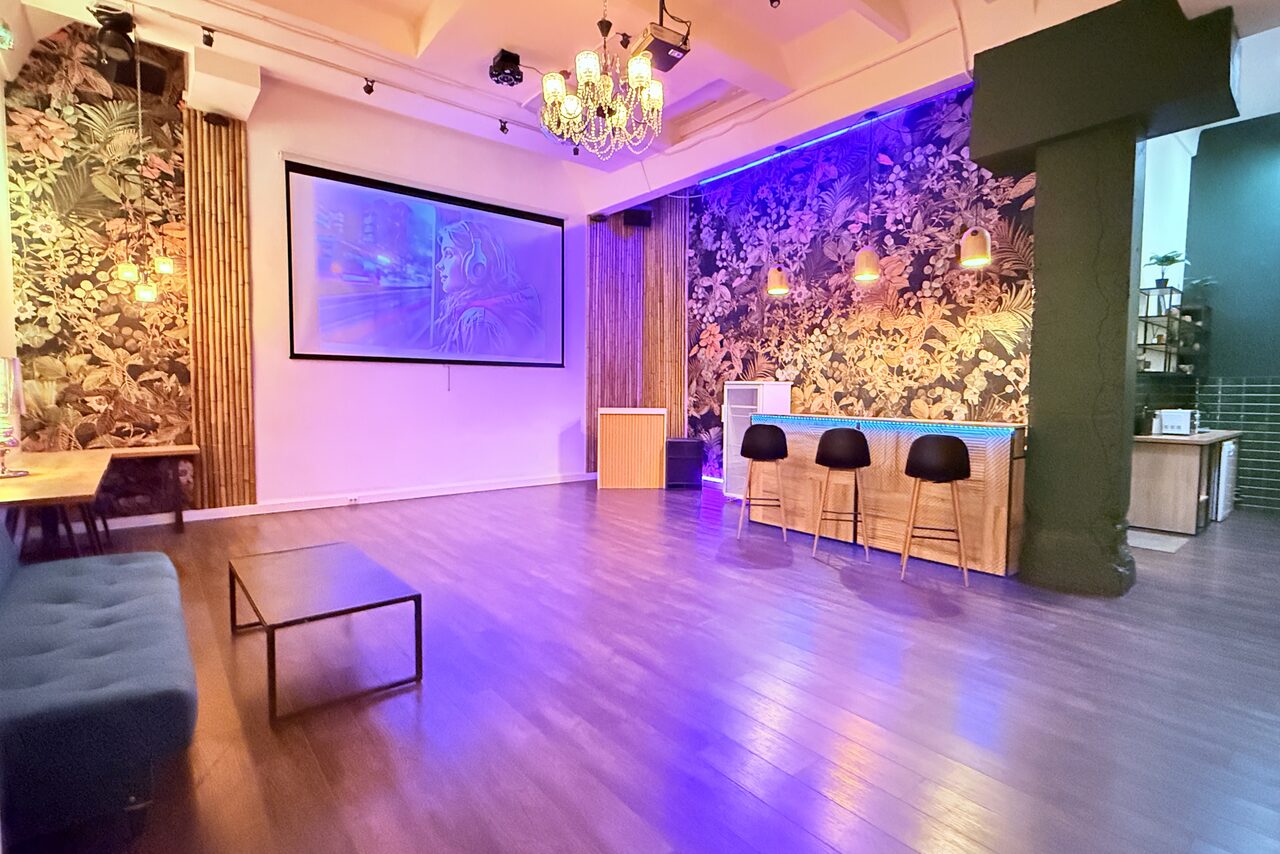 Арендовать Bar on Presnya for private events. Просторное помещение с деревянным полом, барной стойкой с четырьмя стульями, диваном и столами. Стена украшена флористическими обоями. На стене большой экран. Освещение представлено люстрой и светильниками.