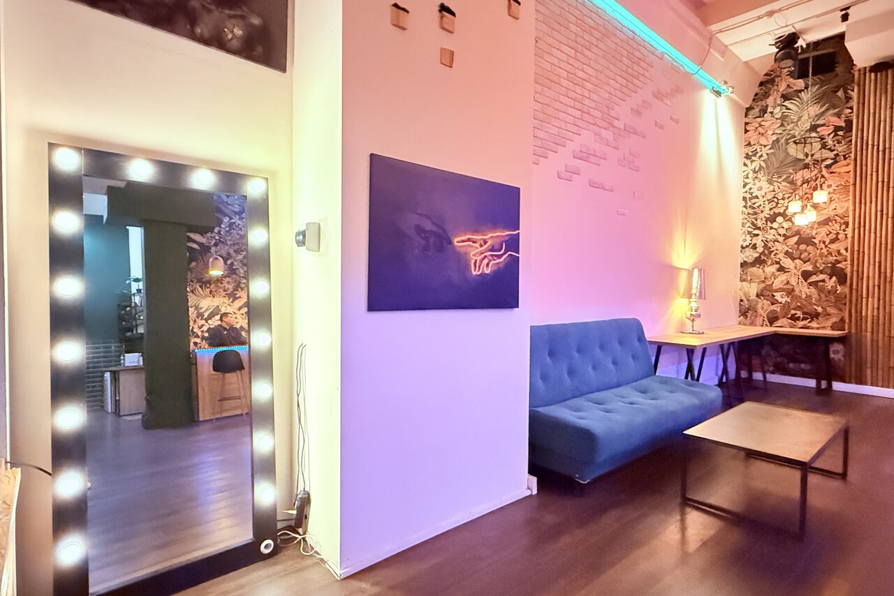 Арендовать Bar on Presnya for private events. Многофункциональное пространство с высоким потолком. Темный пол, пастельные стены, кирпичная кладка. Синяя неоновая подсветка, зеркало с лампочками, мягкий синий диван, низкий столик. На стене декоративное панно и неоновая вывеска.