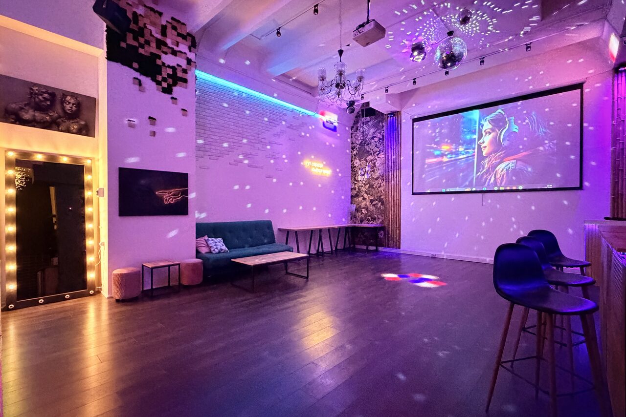 Арендовать Cascad (Каскад). Лофт в центре Москвы с высоким потолком. Пространство с деревянным полом, белыми кирпичными стенами и высоким потолком. В комнате есть диван, столы, стулья, проектор с изображением на экране. На потолке диско-шар и светильники. Украшено разноцветным освещением и рисунком на стене.