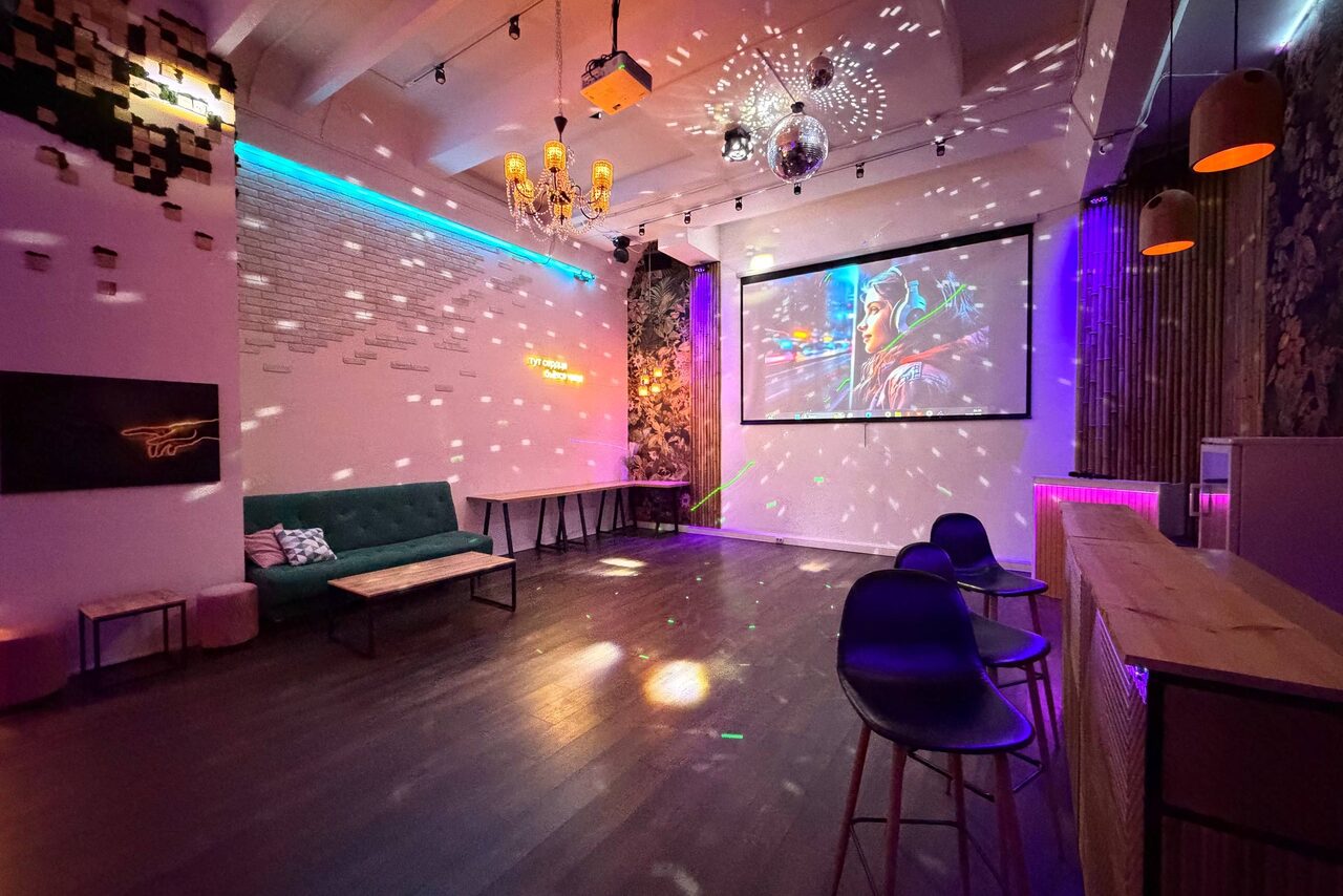 Арендовать Bar on Presnya for private events. Многофункциональное пространство с деревянным полом, барной стойкой и стульями. На стене проектор с изображением, дискотечный шар, разноцветное освещение, диван, журнальный столик, столы. Декор: кирпичная стена, узорчатые обои, подвесные светильники.