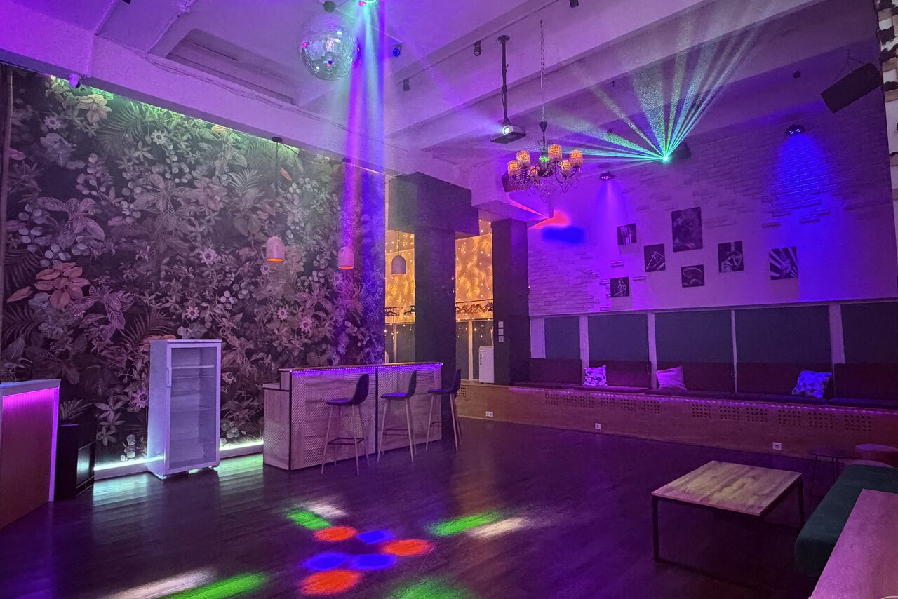 Арендовать Bar on Presnya for private events. Пространство для мероприятий с барной стойкой и двумя барными стульями, диско-шар, разноцветное освещение, столики с диванами у стены. Стена с цветочным принтом, другое с белыми кирпичами и фотографиями. Потолок с открытыми балками.