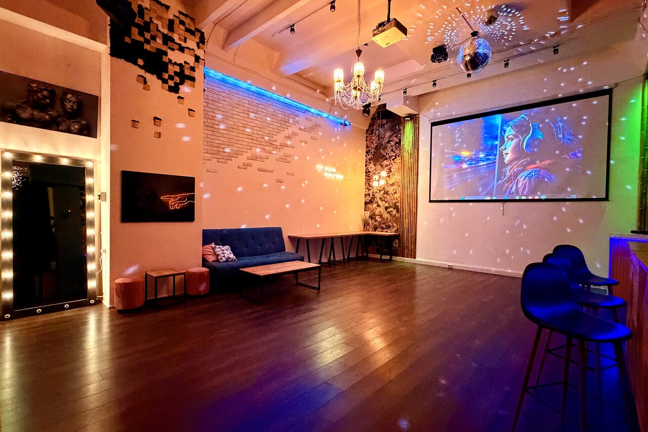 Арендовать Bar on Presnya for private events. Просторное помещение с деревянным полом, синей подсветкой и декоративными элементами. На стене проектор с изображением, зеркальная стена окружена лампочками. Угловой диван, столики и барные стулья. Потолок украшен люстрой и диско-шаром.