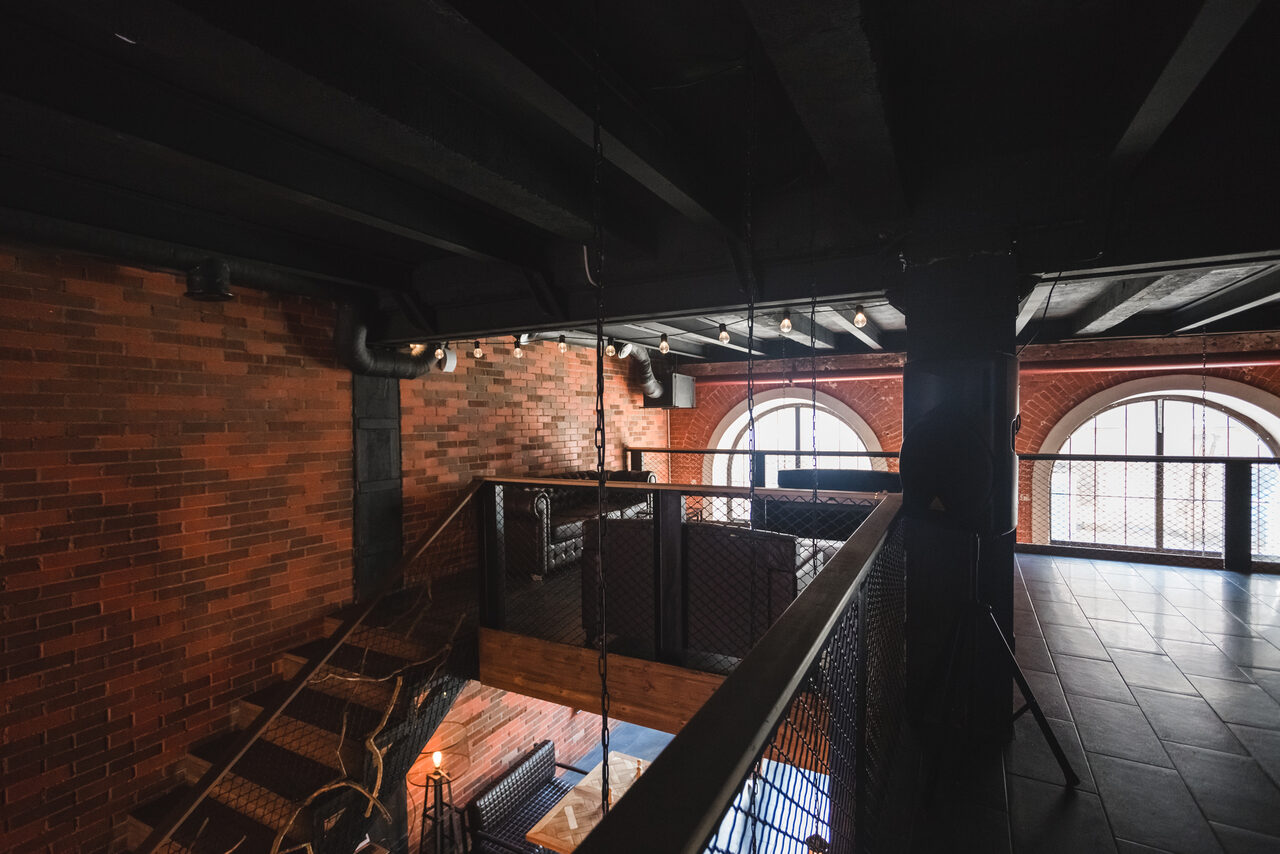 Арендовать Двухуровневый лофт-бар. Лофт-пространство с кирпичными стенами, большим арочным окном и черным металлическим потолком. Лестница из дерева и металла, балкон с металлическими перилами. Подвесное освещение, темная плитка на полу. Обстановка индустриального стиля.