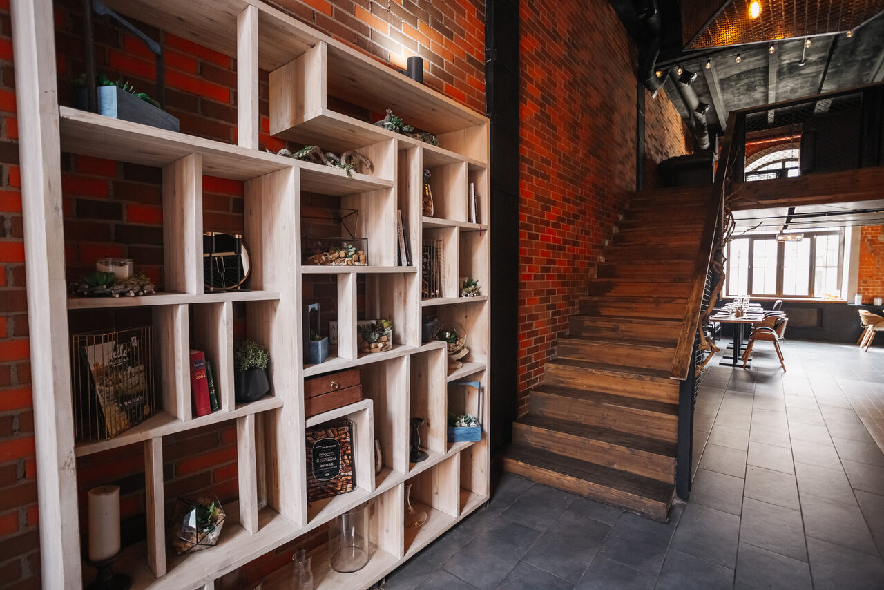 Арендовать Двухуровневый лофт-бар. Лофт-пространство с кирпичными стенами и деревянной лестницей. Справа столы и стулья возле больших окон. Слева стеллаж с декором и растениями. Потолок открытого типа с металлическими элементами. Пол из серой плитки.