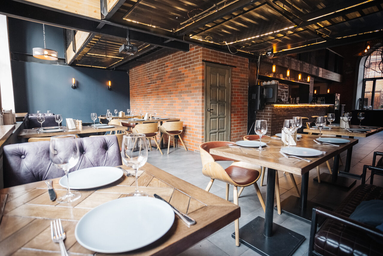 Арендовать Двухуровневый лофт-бар. Современное ресторанное пространство с открытой планировкой; деревянные столы с сервированными бокалами и тарелками; стулья из кожи и дерева; кирпичные стены; металлический потолок; подвесное и настенное освещение; серые и коричневые оттенки.