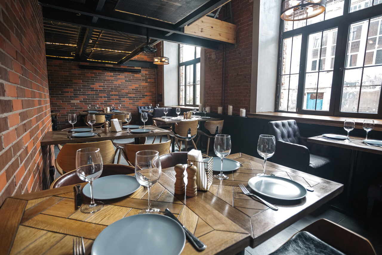 Арендовать Двухуровневый лофт-бар. Ресторанное пространство с кирпичными стенами и деревянным потолком. Столы из дерева с узором, накрытые голубыми тарелками и бокалами. Кожаные стулья, большие окна с черными рамами. Металлические подвесные светильники, направленное освещение.