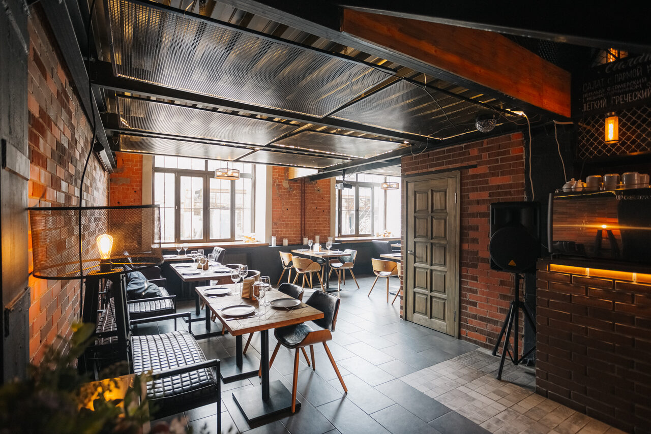 Арендовать Двухуровневый лофт-бар. Пространство для кафе с кирпичными стенами и металлическим потолком. Расставлены деревянные столы и смешанные стулья из дерева и кожи. Большие окна обеспечивают дневное освещение. Потолочные лампы и торшеры добавляют искусственный свет.