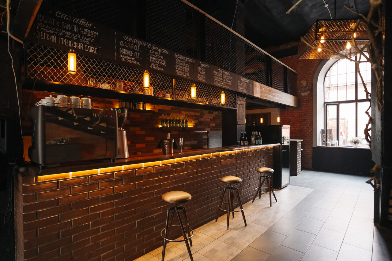 Арендовать Двухуровневый лофт-бар. Барная зона с кирпичной стойкой и тремя табуретами, черные сетчатые перегородки, коричневая плитка, подвесные светильники с теплым светом, большая арка с окнами, меню написано мелом на досках, металлические элементы интерьера, стены из красного кирпича.