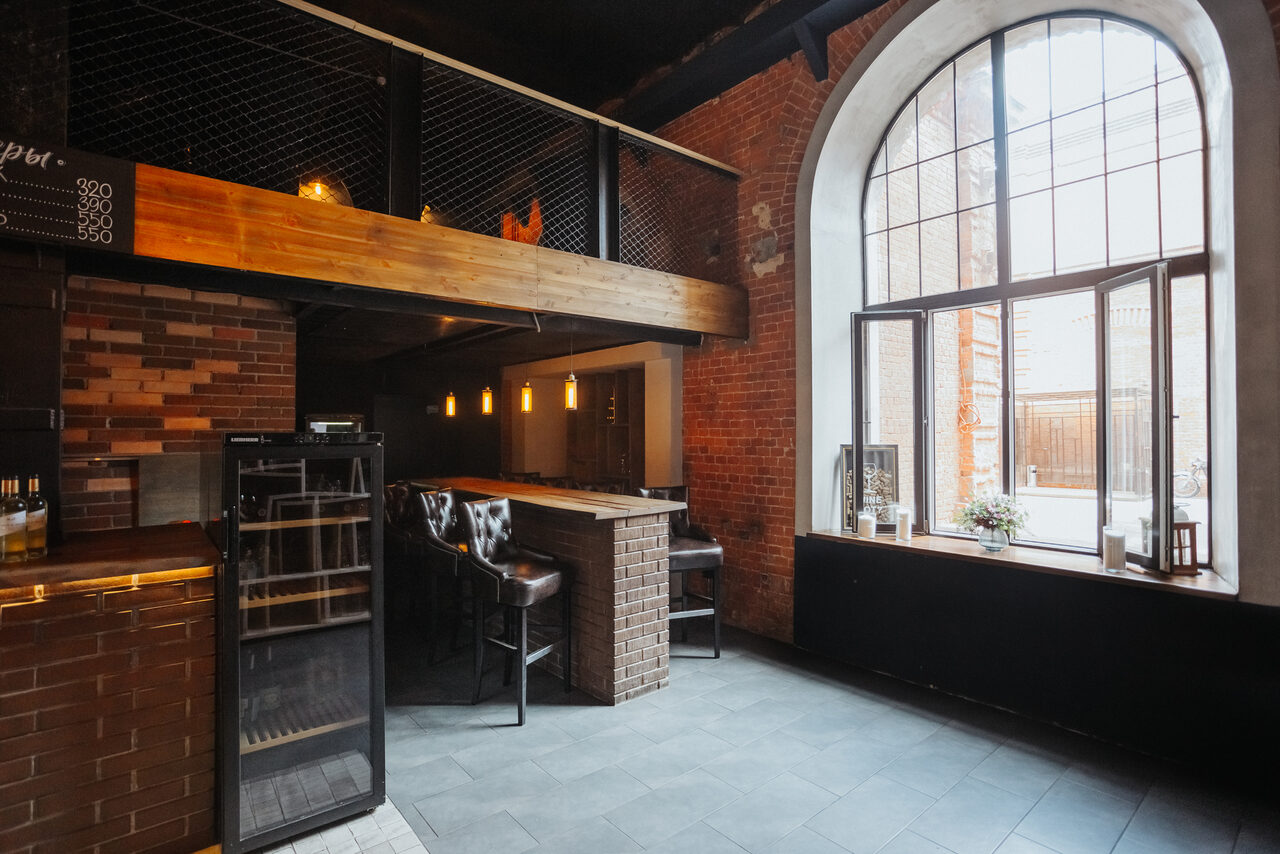 Арендовать Двухуровневый лофт-бар. Лофт пространство со столиком барного типа и высокими стульями, кирпичные стены, большой арочный оконный проем, светильники подвесного типа, пол серого цвета, барная стойка обложена плиткой, металлические и деревянные элементы в отделке.
