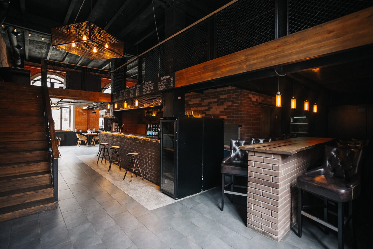 Арендовать Двухуровневый лофт-бар. Пространство в стиле лофт с кирпичными стенами и темными деревянными элементами. Два уровня с лестницей. Барная стойка с высокими стульями. Подвесное освещение, пол из серой плитки, черный потолок. Столы и стулья в зоне отдыха.