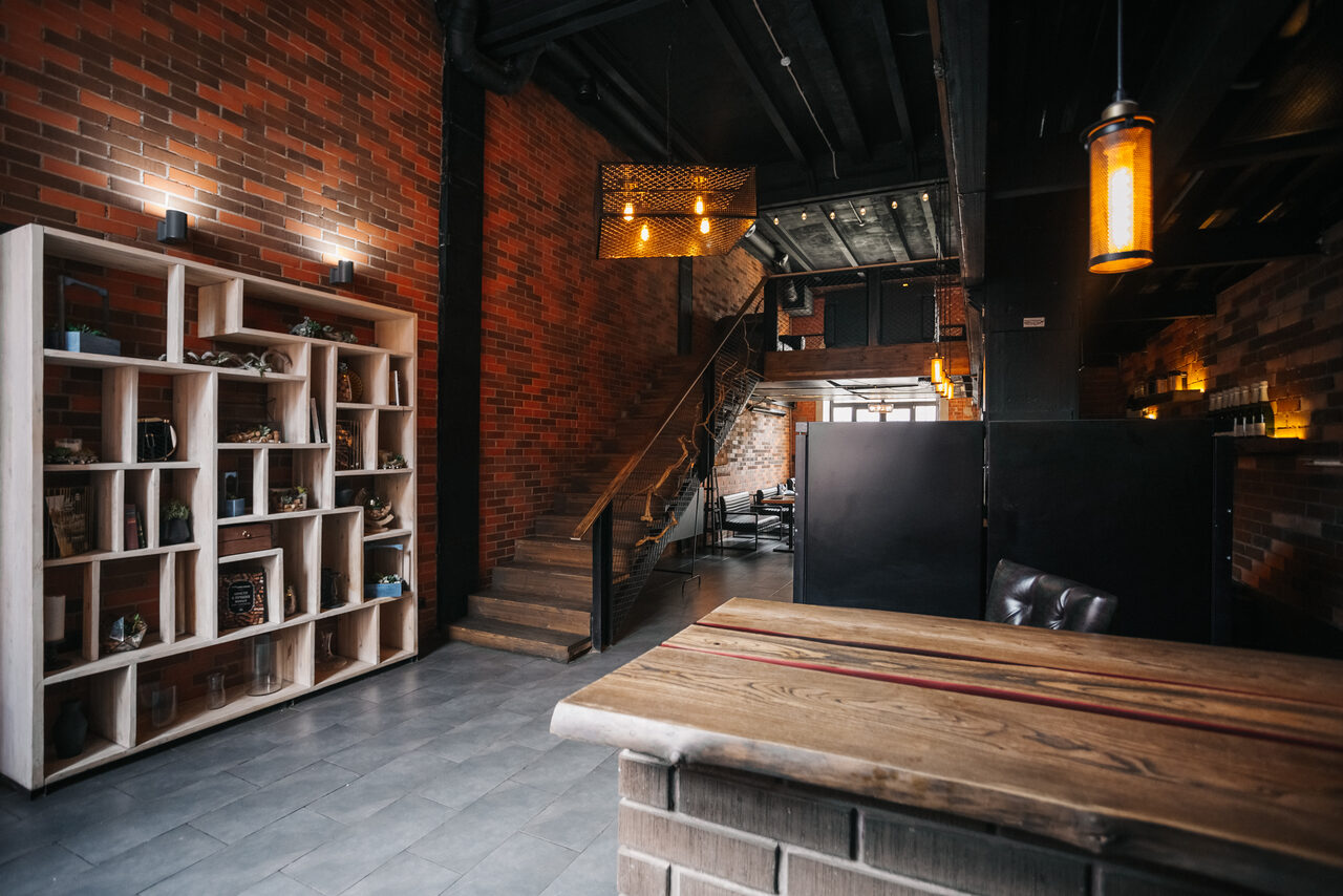 Арендовать Двухуровневый лофт-бар. Лофт-пространство с кирпичными стенами и деревянной меблировкой. Видна лестница, ведущая на второй уровень, деревянный стол перед стеллажом с полками, кресло. Освещение подвесными лампами, теплый свет. Пол из серой плитки.