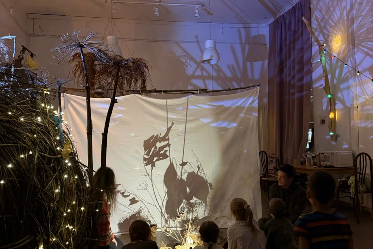 Арендовать Children's eco-loft. Комната с белыми стенами, натяжным потолком и подвесными светильниками. На переднем плане белый экран с теневым театром. Справа стол с предметами, стулья. Декорации из веток и гирлянды с лампочками. На полу сидят дети.