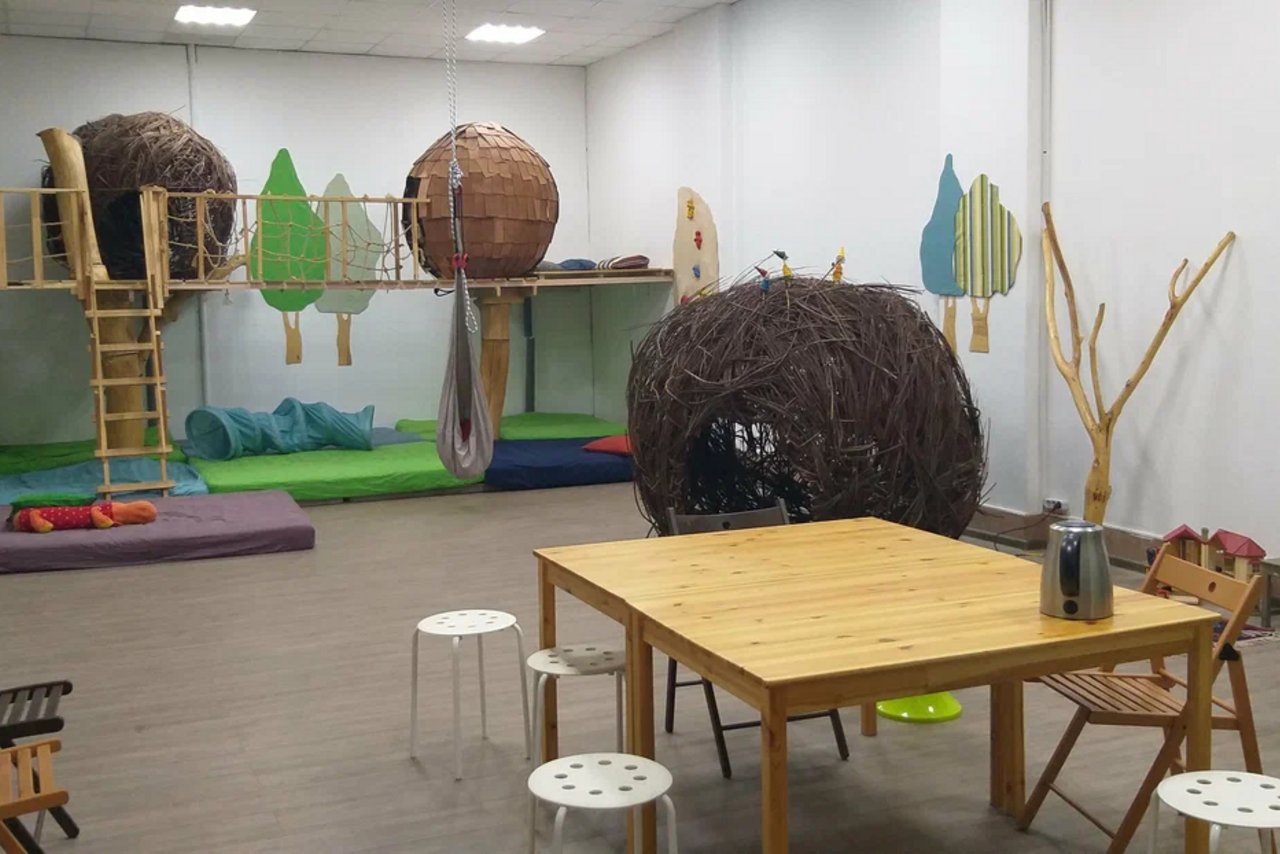 Арендовать Children's eco-loft. Детская игровая комната с деревянными конструкциями, имитирующими гнёзда. Лестница, подвесной гамак, стол, стулья, мягкие подушки и коврики. Светлые стены с нарисованными деревьями. Потолочные светильники. Пол из деревянных панелей.