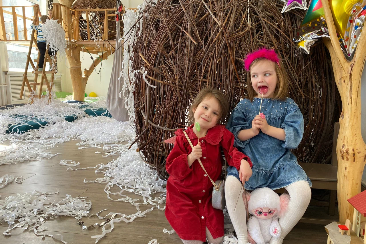 Арендовать Children's eco-loft. Изображено детское игровое пространство с деревянными элементами и конструкцией из веток. Пол покрыт светлыми панелями, вокруг разбросаны белые бумажные ленты. Яркое естественное освещение, на стене окна. Детская игрушка и разноцветные воздушные шары.