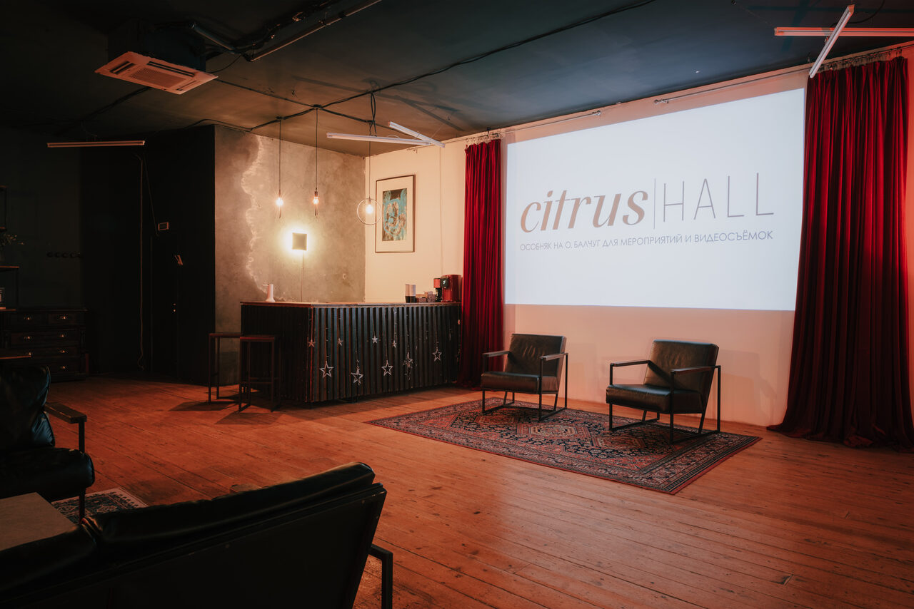 Арендовать Citrus Hall (Цитрус Холл). Лофт Грейпфрут. Просторное помещение с деревянным полом, черным потолком и красными занавесками. На стене крупный экран с надписью. Две черные кожаные кресла на ковре. Справа барная стойка. Освещение от подвесных ламп и светильников на стене.