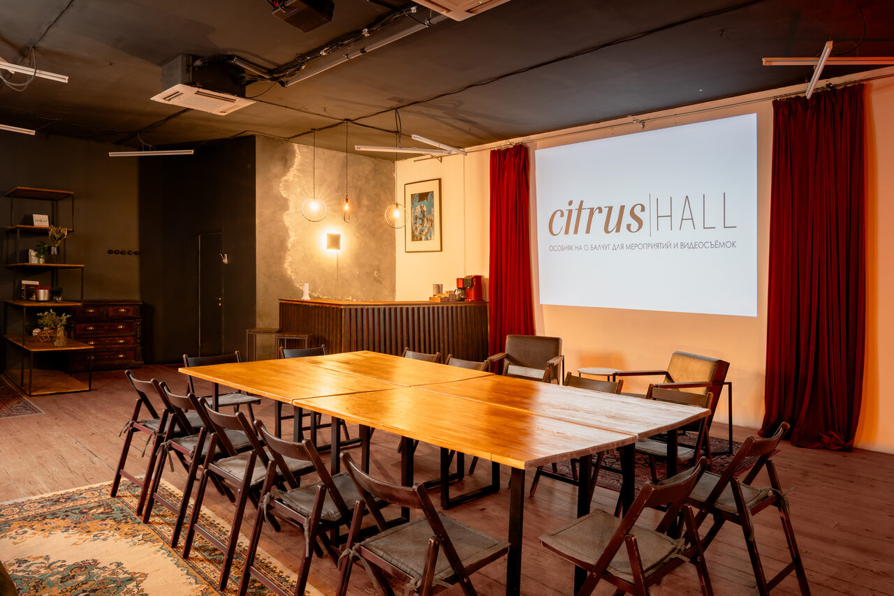 Арендовать Citrus Hall (Цитрус Холл). Лофт Грейпфрут. Креативное пространство с деревянными столом и складными стульями. На стене проектор с изображением "citrus HALL". Темные и светлые стены, красные шторы. На полу ковер, с левой стороны полки и комод. Потолочное освещение и декорации.