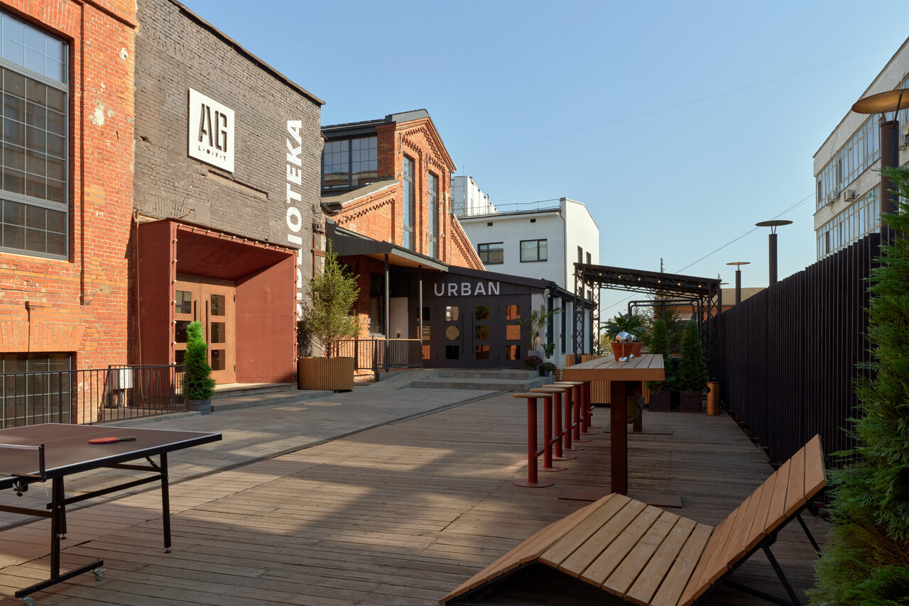 Арендовать Spacious loft with a vintage car. Площадка на открытом воздухе с деревянным настилом, длинными деревянными скамейками и столами, стол для пинг-понга. Здание из красного кирпича с большими окнами. Вывески "AG" и "URBAN". Планировка открытая, высокие стены. Растения в горшках.