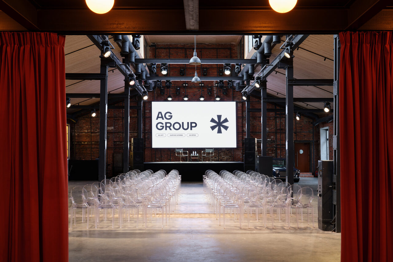 Арендовать Spacious loft with a vintage car. Большое помещение с высокими потолками, обставленное прозрачными стульями, расположенными рядами. На сцене установлен экран с надписью «AG GROUP». Стальные балки и прожекторы на потолке. Стены кирпичные, свет от подвесных ламп. Шторы красные.