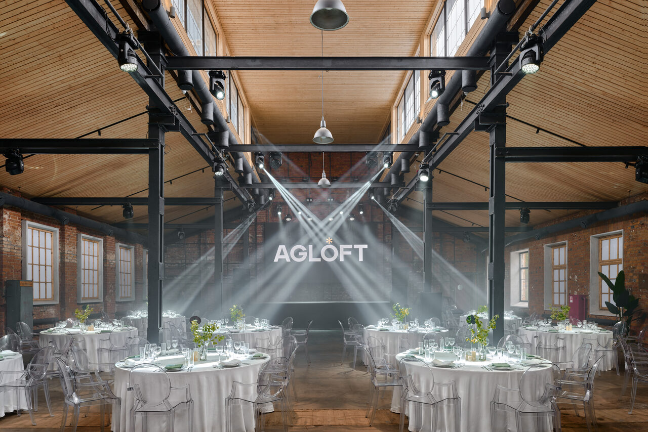 Арендовать Spacious loft with a vintage car. Лофт-пространство с высокими деревянными потолками и кирпичными стенами. Размещены круглые столы с белыми скатертями и прозрачными стульями. Простор освещается большими окнами и подвесными светильниками. На задней стене надпись «AGLOFT». Помещение в хорошем состоянии.