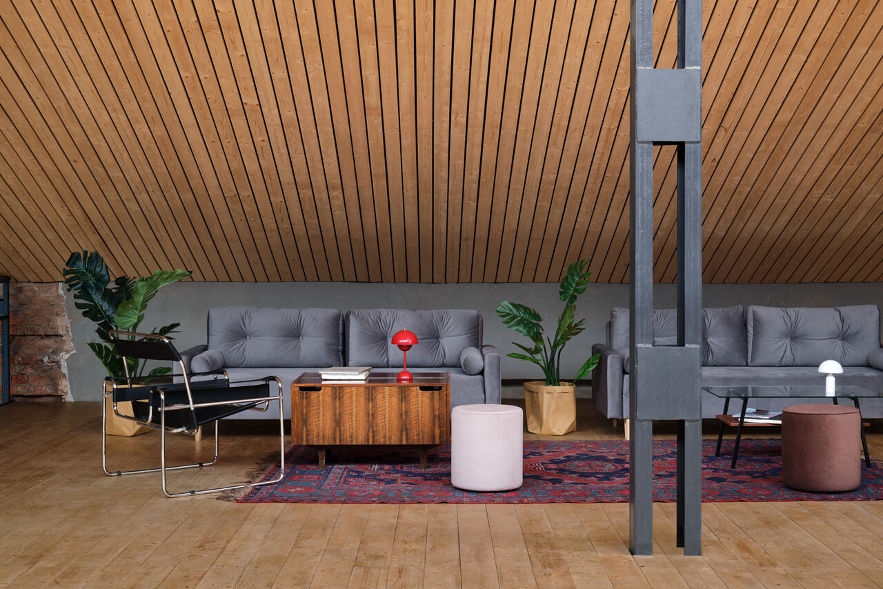 Арендовать Spacious loft with a vintage car. Чердачное пространство с косым деревянным потолком и деревянным полом. Две серые диваны, кресло, журнальный столик, ковёр и два пуфа. Рядом комнатные растения в горшках. Освещение с красной и белой настольными лампами.