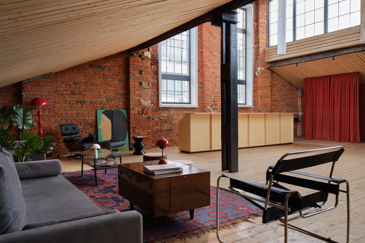 Арендовать Spacious loft with a vintage car. Лофт-пространство с кирпичными стенами, деревянным полом и наклонным потолком. Мебель: диван, кресла, деревянный стол, ковер. Большие окна, естественное освещение. Красные шторы, растение. Металлические и деревянные элементы. Декорирована лампа, книги на столе.