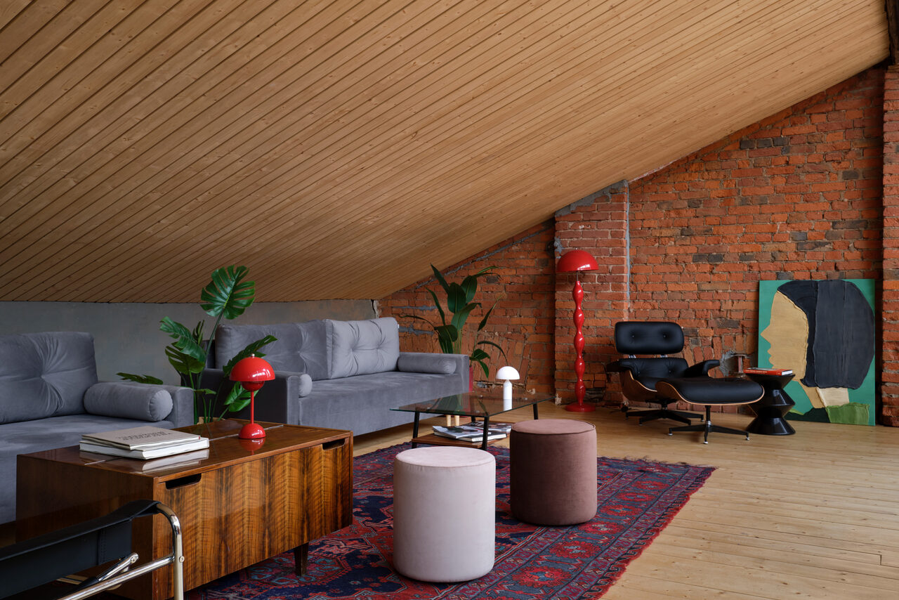 Арендовать AG Loft. Urban (АГ Лофт. Урбан). Просторный лофт с раритетным автомобилем. Лофт с наклонным деревянным потолком и кирпичными стенами. Серый диван, деревянный столик, кресло, пуфы, ковёр с орнаментом. Настольные лампы красного цвета, комнатные растения. Настенное искусство, светлый деревянный пол.