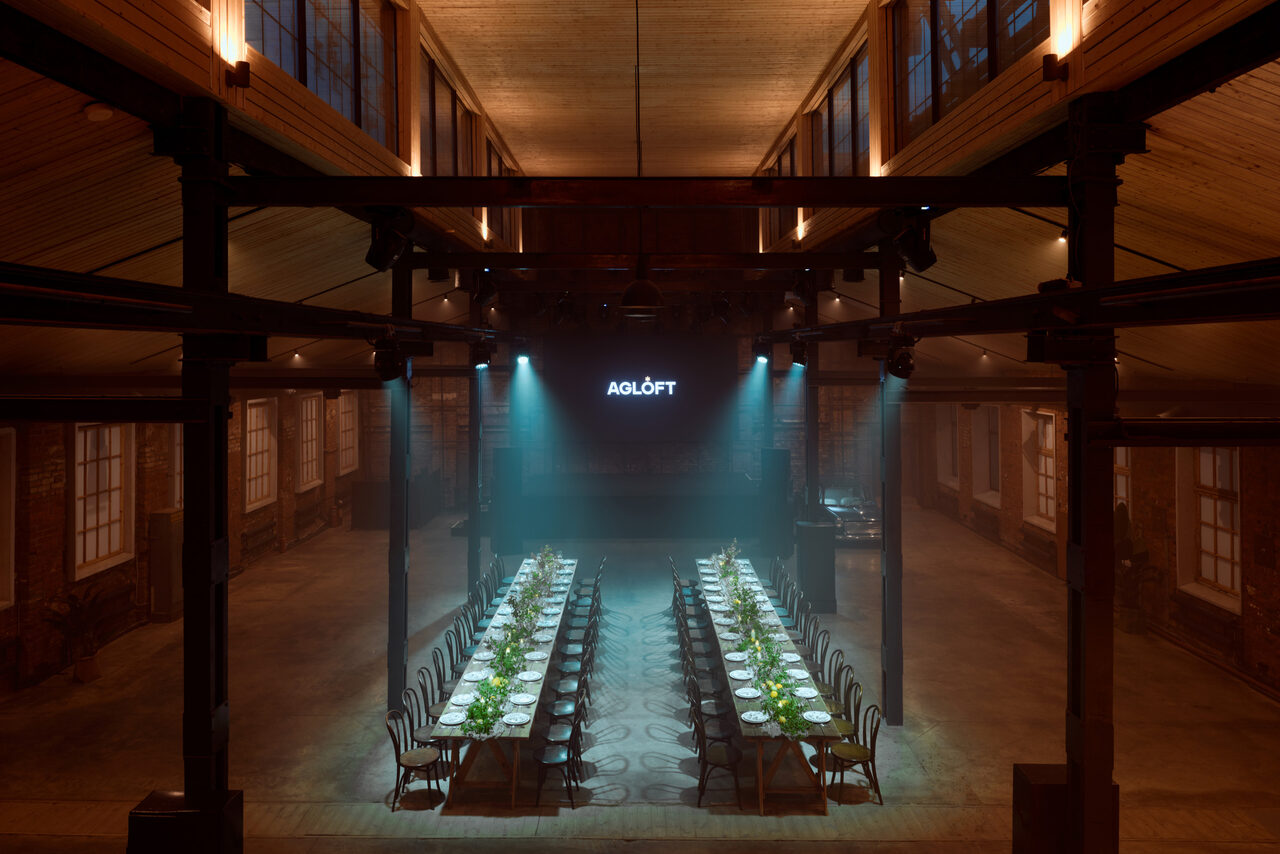 Арендовать Spacious loft with a vintage car. Просторное помещение с высокими потолками, деревянными элементами и кирпичными стенами. Длинные столы, выстроенные в два ряда, накрыты посудой и украшены зелёными растениями. На стене дисплей с надписью "AGLOFT". Мягкое освещение сверху.