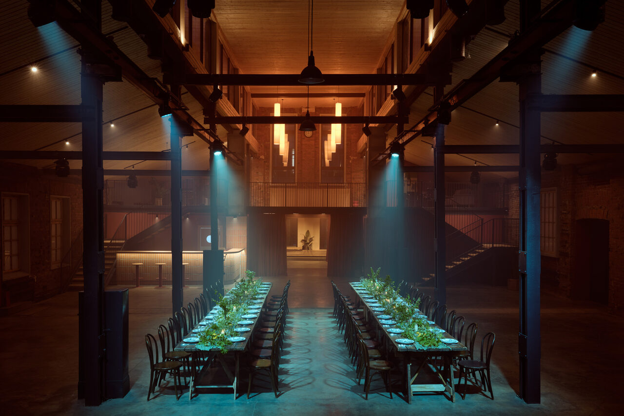 Арендовать Spacious loft with a vintage car. Просторное помещение с высокими потолками, деревянные балки, две длинные сервированные столы с черными стульями, лестницы по бокам, направленное светодиодное и потолочное освещение, кирпичные стены, декор из растений на столах, уютная атмосфера.