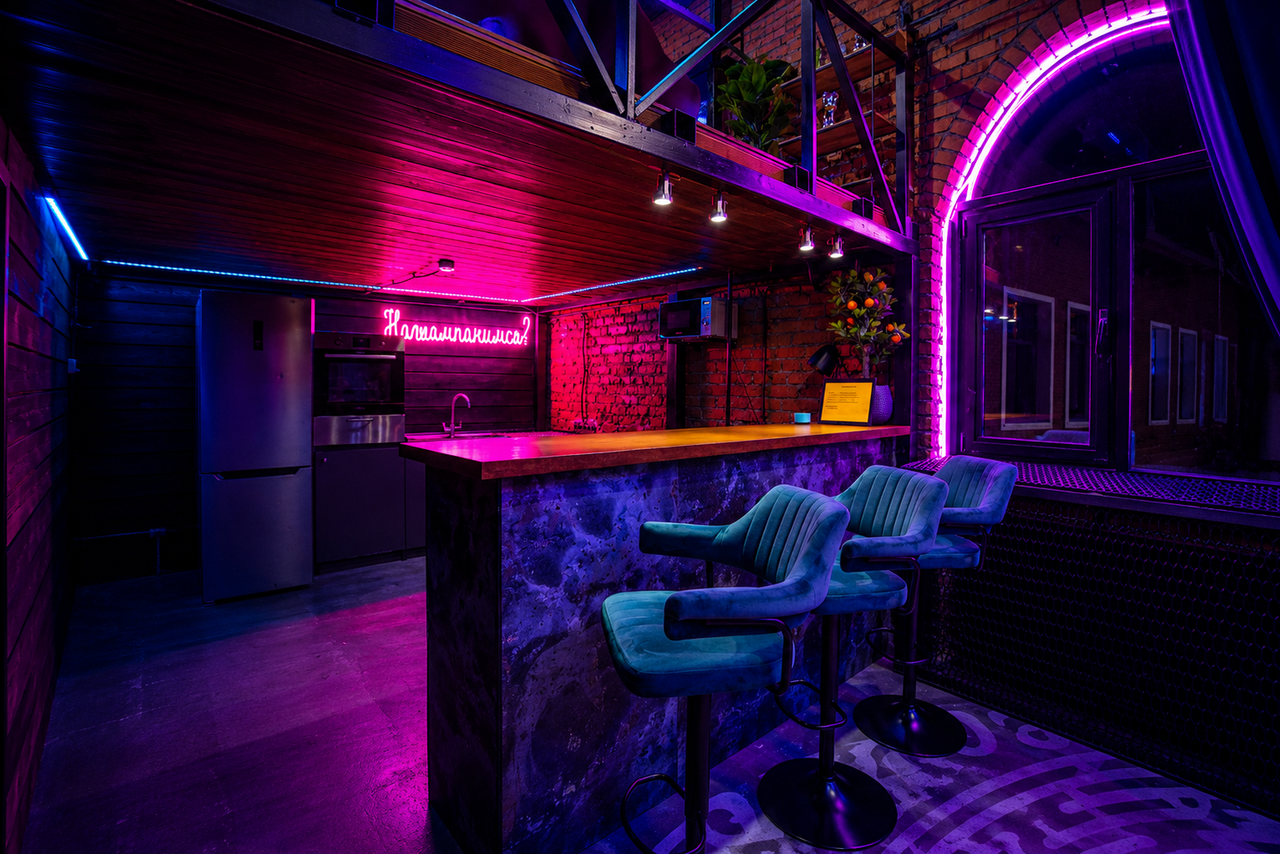 Арендовать Duplex loft in the art space . Барное пространство с деревянной стойкой, барными стульями и холодильником. Кирпичные стены, неоновая подсветка, арочное окно с занавеской, деревянный потолок. Светодиодная вывеска и цветы на стойке.