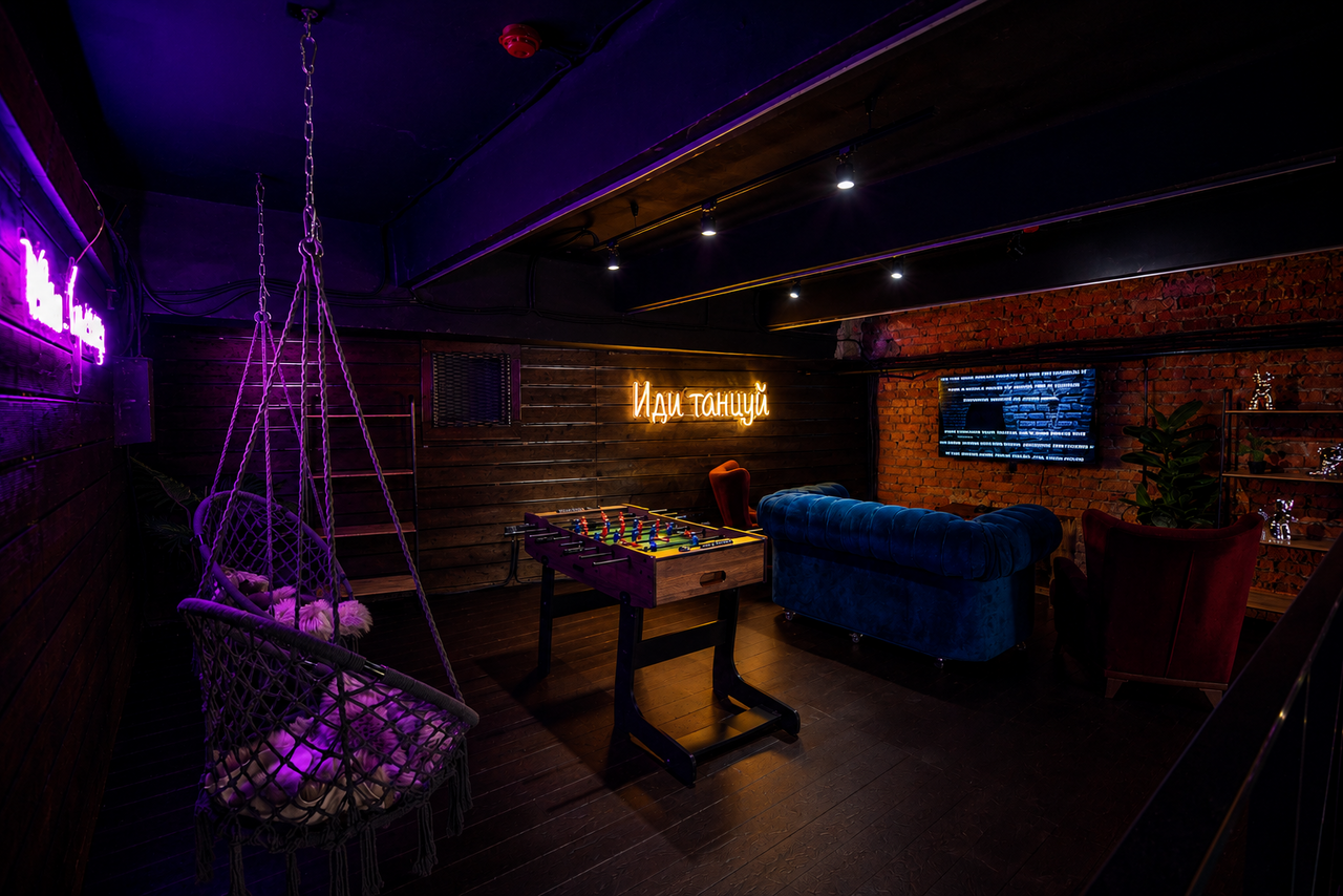 Арендовать Duplex loft in the art space . Лаунж-зона с настольным футболом, креслами, подвесным креслом и диваном. На стенах неоновые надписи на русском и телевизор. Темное освещение, деревянные и кирпичные стены, пол из темного дерева.