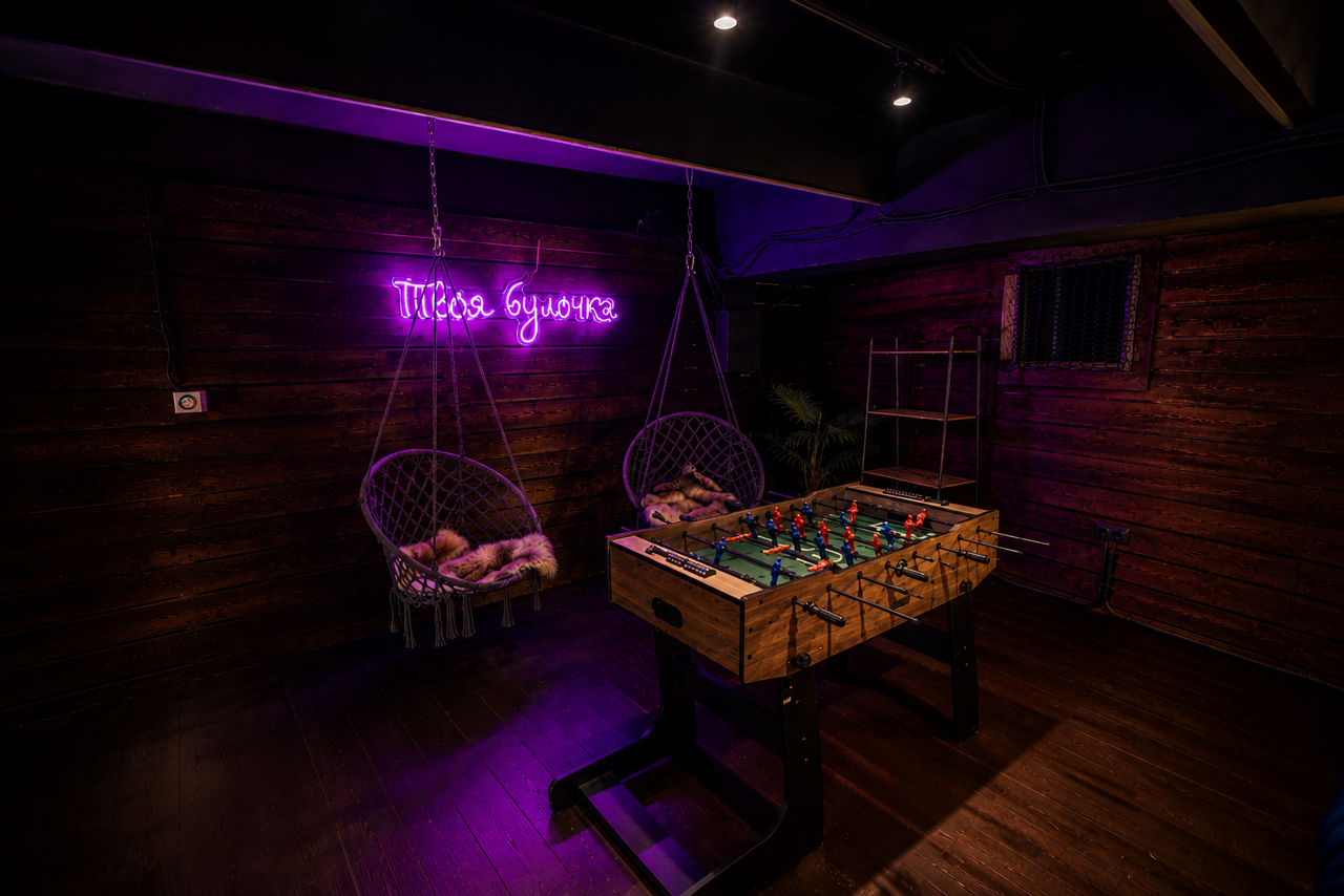 Арендовать Duplex loft in the art space . Темное помещение с деревянными стенами и полом. В центре находится футболный стол. Справа подвесные кресла с меховыми подушками. На стене висит неоновая надпись на русском языке. Освещение точечное, фиолетовый и теплый свет.