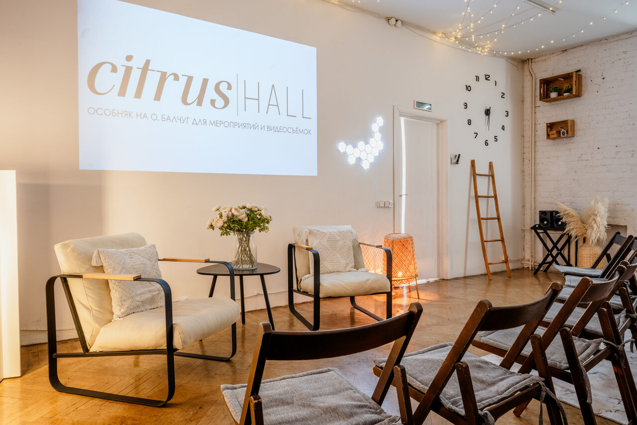 Арендовать Citrus Hall (Цитрус Холл). Зал Свити. Просторное светлое помещение с паркетным полом и белыми кирпичными стенами. Ряды деревянных стульев, кресла с подушками, стеклянный столик с вазой, настенные часы, лестница, проектор с изображением. Потолочные гирлянды и боковое освещение.