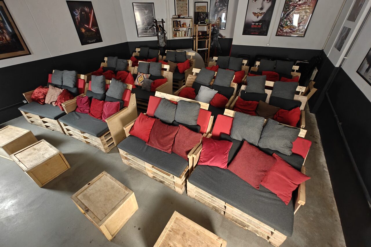 Арендовать Chamber cinema. Домашний кинотеатр с самодельными креслами из деревянных поддонов, серыми и красными подушками. Стены украшают постеры. Освещение приглушенное. На полу серый пол, перед креслами два небольших деревянных стола.