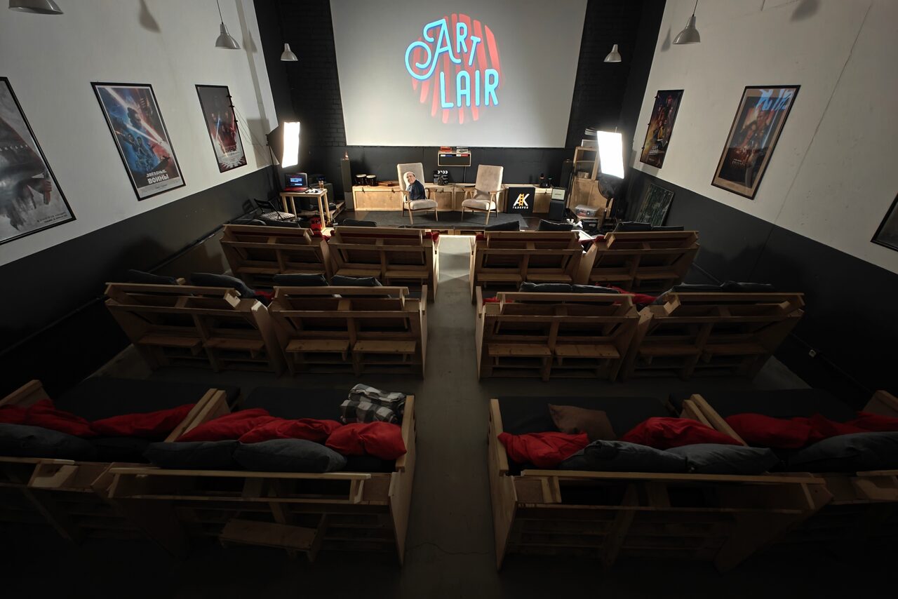 Арендовать Chamber cinema. Пространство в виде зала с проектором и экраном, который показывает логотип "Art Lair". Ряды деревянных кресел с подушками и пледами. По бокам стены украшены плакатами. Нейтральные тона, рассеянное освещение.